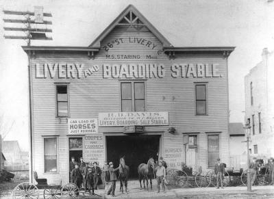 Two story wood structure with wagons, horses and men out front.
