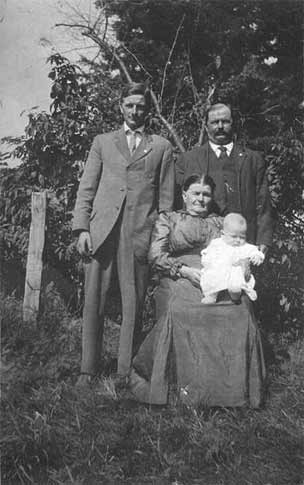 Susan seated in the pasture with baby in arms and son and granson behind the chair.