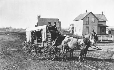 Two guys on a stagecoach with two horses and buildings in background.